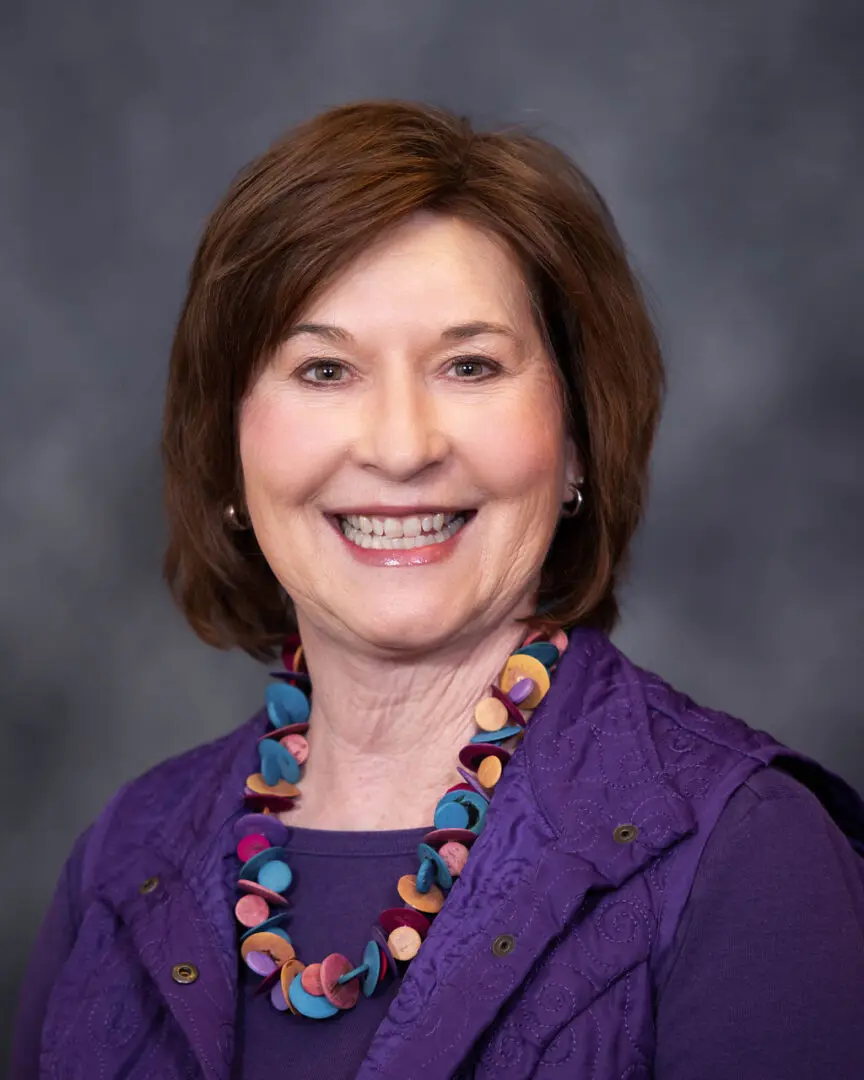 A woman in purple shirt and necklace smiling.